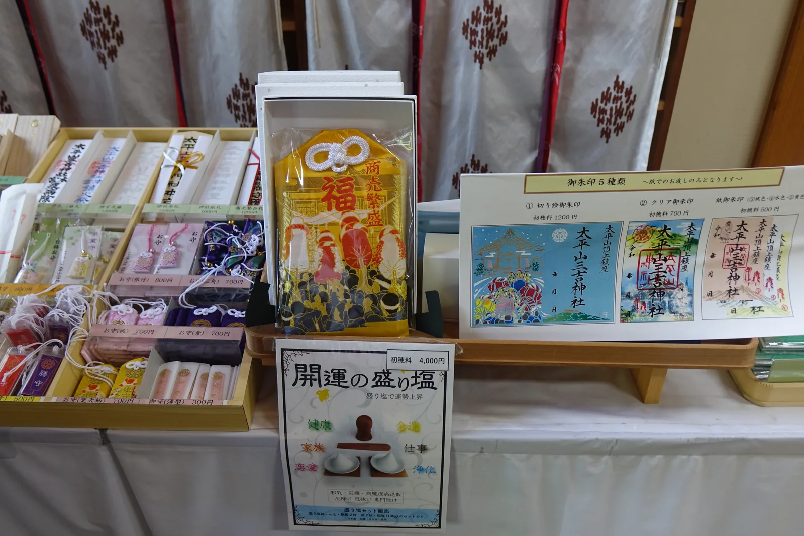授与所的桌面。御守分格擺開，中央立著「開運の盛り塩」的說明牌，初穗料（hatsuhoryō，接受授与品時獻納的金額）四千日圓；右邊是五種御朱印（goshuin，參拜後由神社書寫社名與日期的朱印）的樣張。背後的白幕印著神紋。