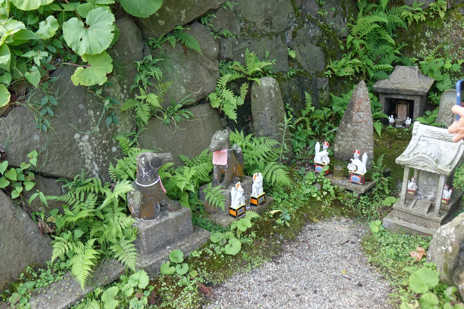 石垣腳下一排石祠（sekishi，石造的小型祠堂）與石像。狐狸形的石像有好幾尊，大小不一，中間夾著石塔與小型的石燈籠，周圍長滿蕨與苔。