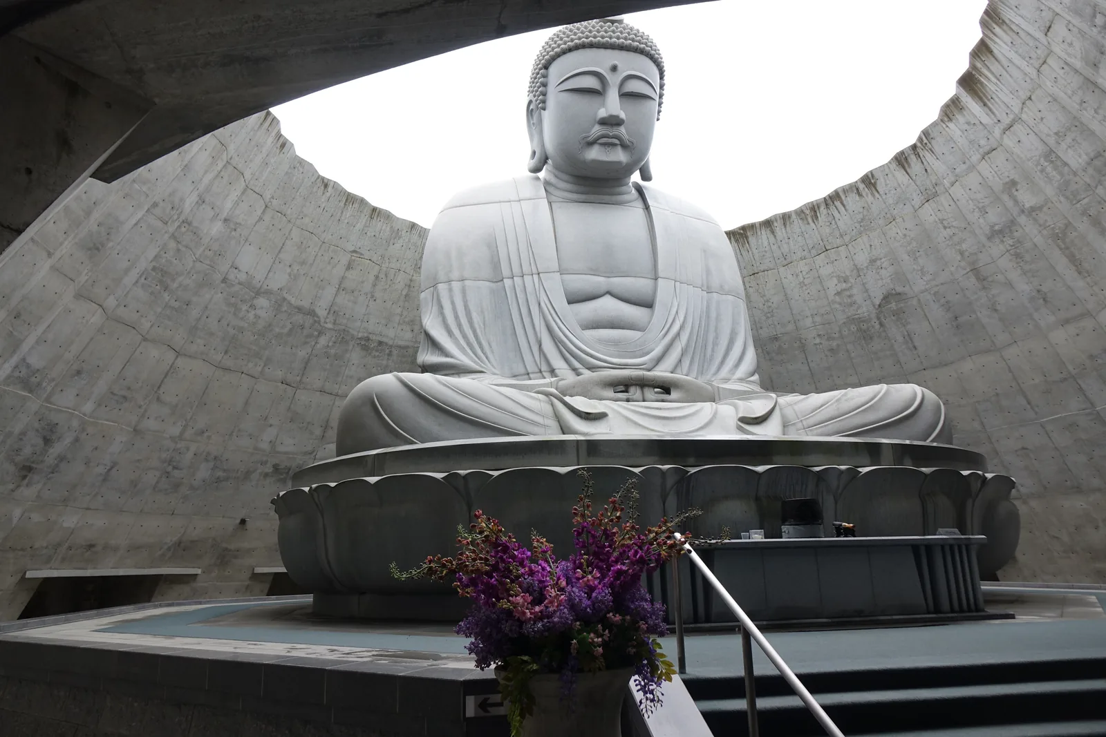 頭大佛的石造蓮座與座前的紫色供花與黑色香台，穹頂內側可見放射狀的混凝土肋