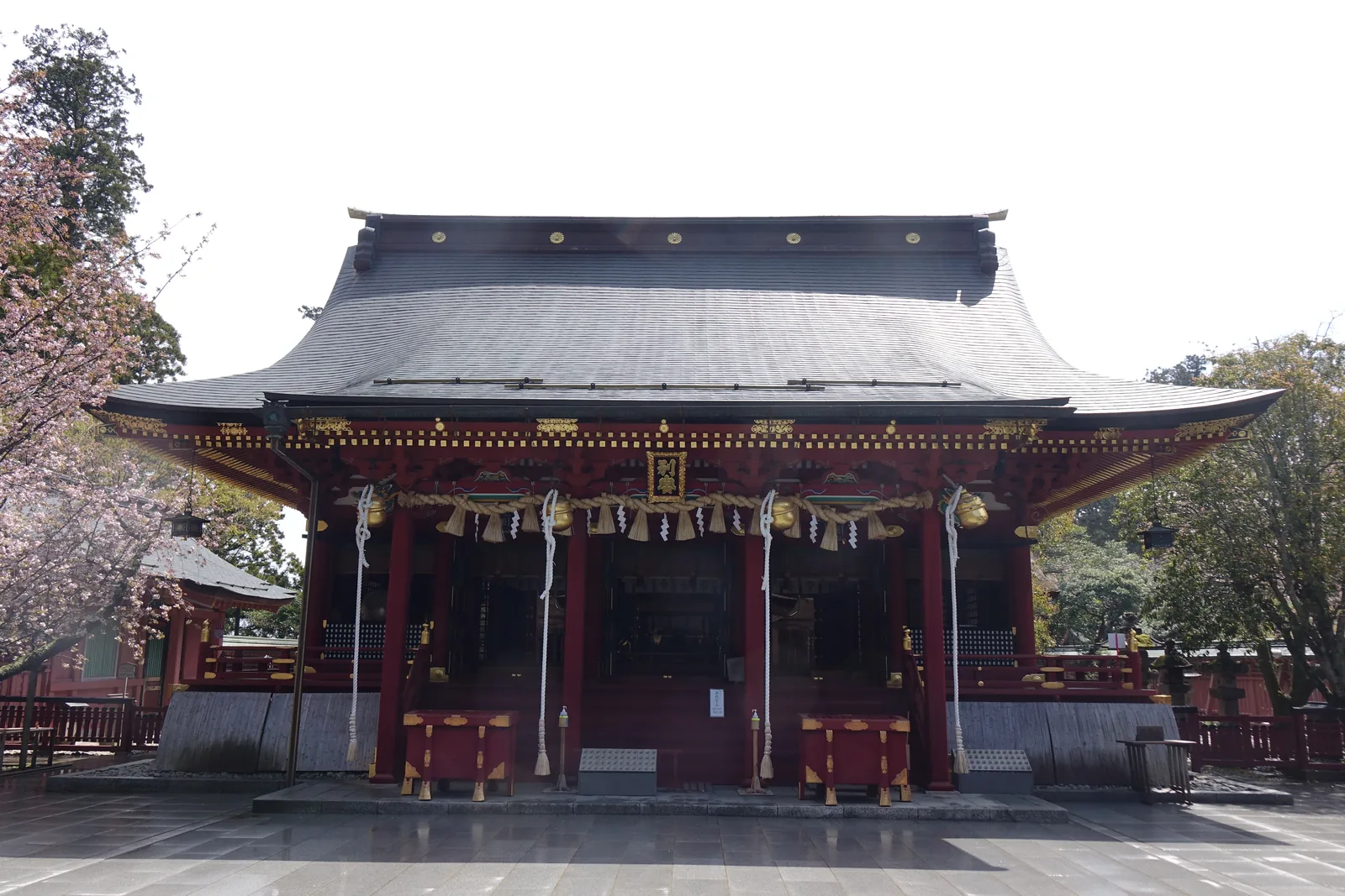 拜殿的正面。淺灰色的大屋頂在中央高起，屋簷向兩端舒展；柱與欄杆是朱色，簷下橫著注連繩與五個金色的鈴。左右各接出一段朱色的迴廊。地面積著雨水，映出建築的倒影。
