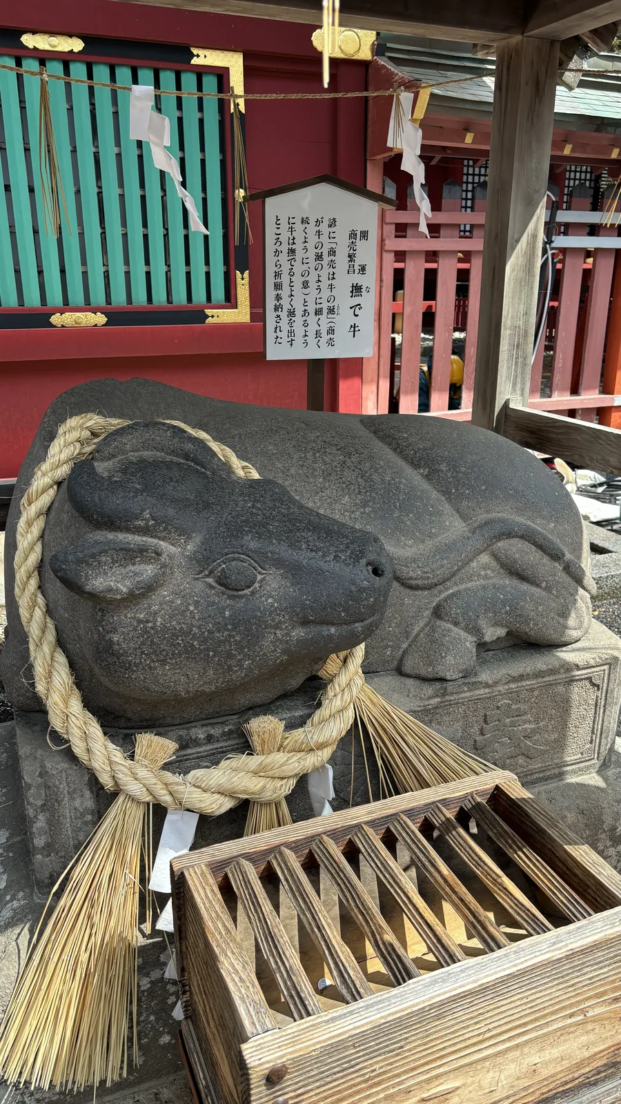 一頭石造的臥牛伏在石台座上，脖子上掛著粗繩與稻草束，牛角向後彎。台座正面刻著「牛」字。後方的立牌題「開運　商売繁昌　撫で牛」，說明牛被撫摸就會流涎，因而以「商売は牛の涎」的諺語被奉納於此。背景是朱色與綠色的社殿。