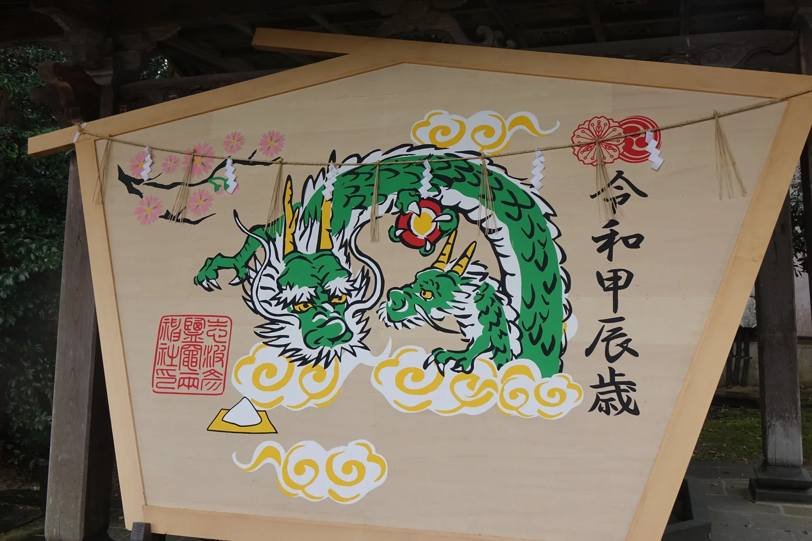 一面素木的大繪馬靠在簷下，上緣拉著繩子與紙垂。畫面中央是兩條相對的綠色龍，周圍是黃白色的雲，左上角一枝粉色的櫻，右側直書「令和甲辰歲」。左下角壓著「志波彦神社鹽竈神社」的朱印，朱印下方畫著一小堆盛在黃色台上的白鹽。