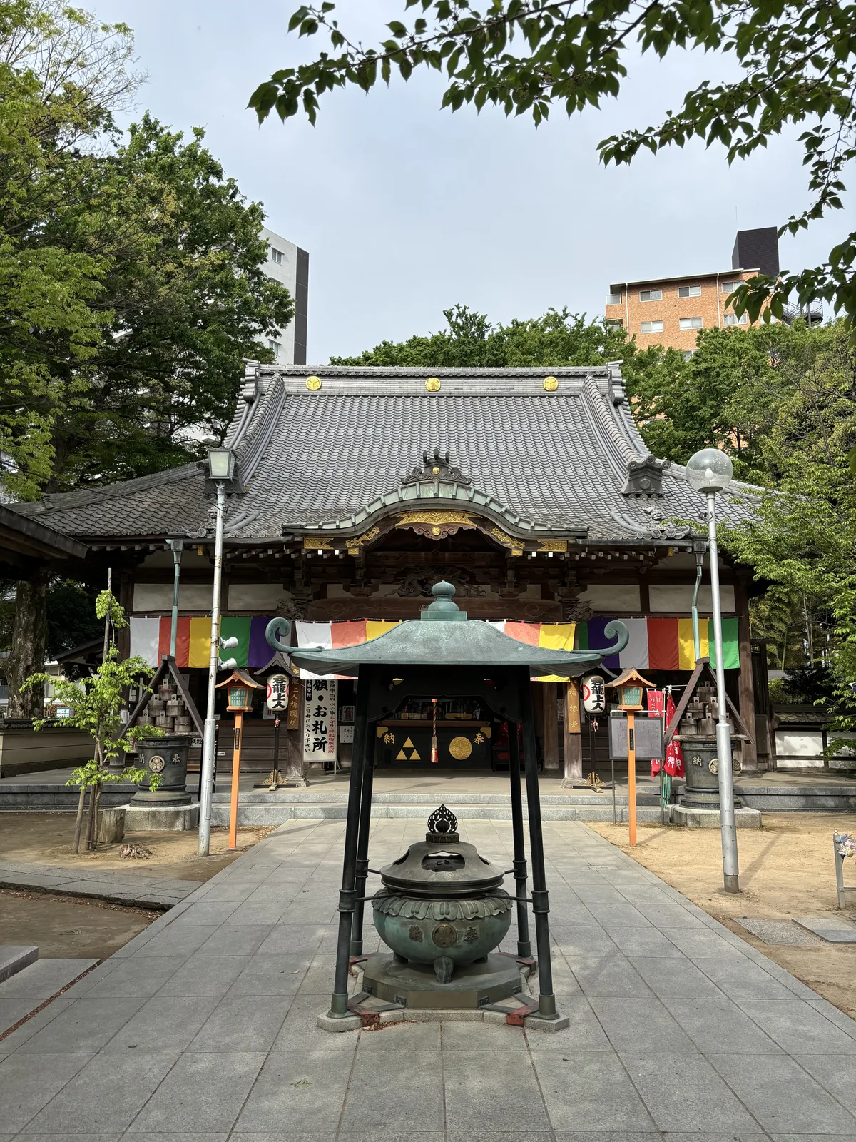 同一棟堂的全景。前景一只銅製的常香爐，上方另加一頂青綠色的小屋頂，四根細鐵柱撐著。堂的左右兩側各立一座木造的燈籠與一只奉納木桶。屋脊上三枚金色的圓紋，背後探出後方的公寓。