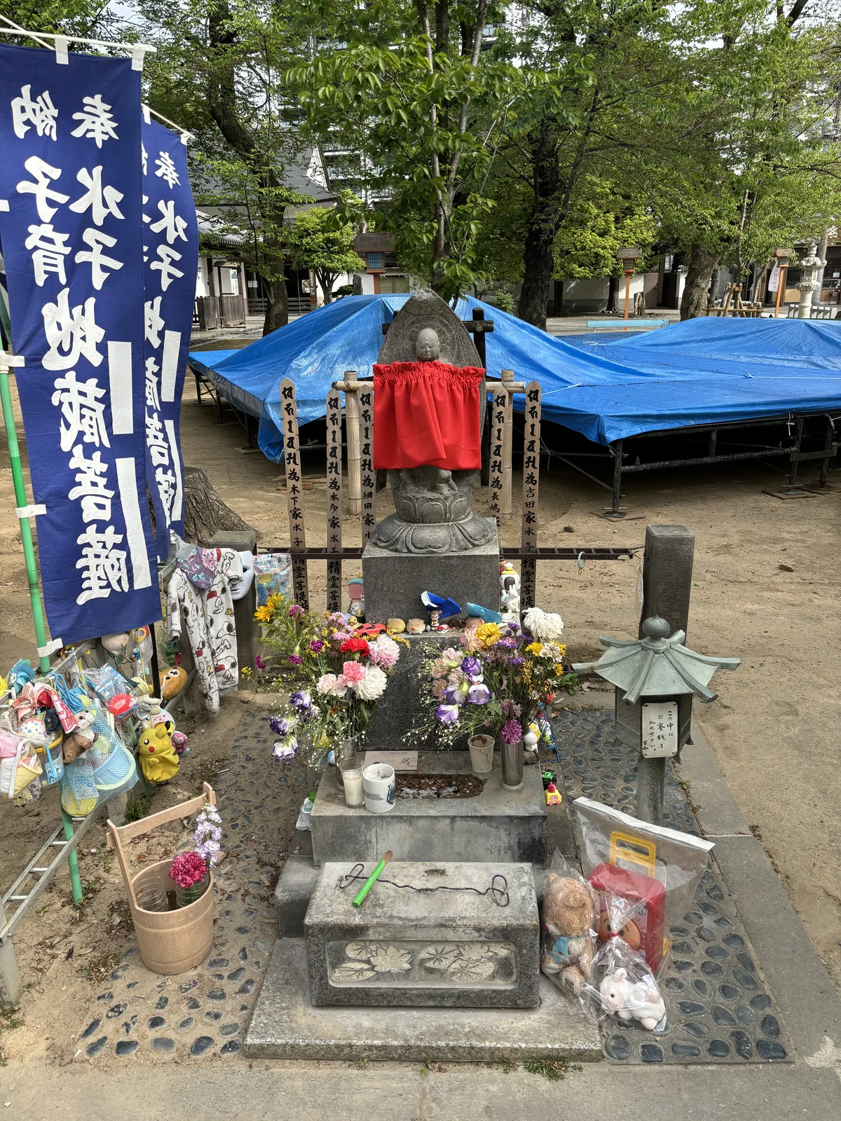 石造的地藏坐像，戴紅色圍兜，背後一排竹製卒塔婆寫「南無阿彌陀佛　為某某家水子精靈菩提」，讀得出本下、本多、細田、長嶋、吉田五個家名。腳邊堆著毛絨玩偶、皮卡丘、玩具車與紙風車，一層疊一層；右側木桶裡插著新的康乃馨。左方藍幟寫「奉納　水子育兒地藏菩薩」。後方那片藍色帆布蓋的是收起來的攤架。