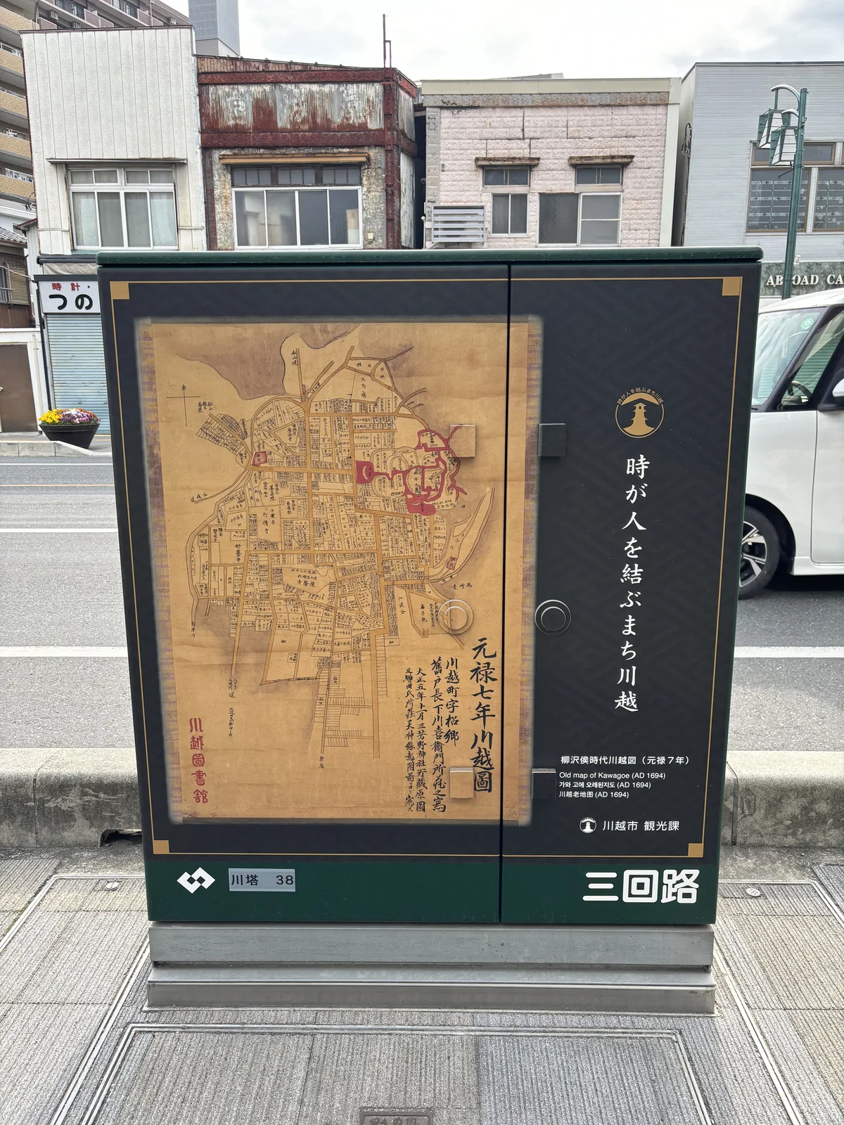 人行道上的電線共同溝地上機器，箱門印著一張褐色的古地圖，右下角直書「元祿七年川越圖」，落款「川越圖書館」。右半黑底白字寫「時が人を結ぶまち川越」，意思是「時間把人繫在一起的城市，川越」；下方標註「柳澤侯時代川越圖（元祿七年）／Old map of Kawagoe (AD 1694)」，發行者川越市觀光課。箱底編號「川塔 38」。背後是一整排看板建築。