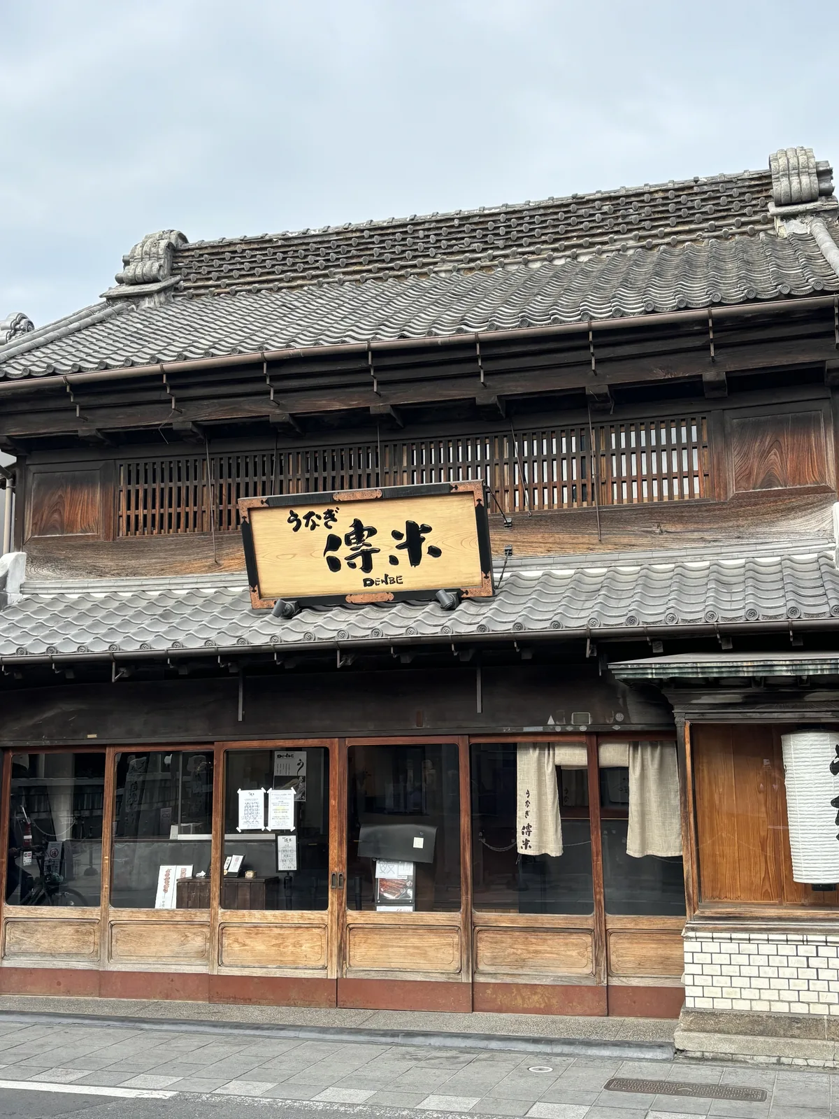 同一棟從街對面看。灰色的屋瓦、黑色的木格子、白色的漆喰（しっくい，shikkui，石灰抹面）腰壁，招牌懸在二樓與一樓屋簷之間。左端另一棟蔵造り的商家，鬼瓦探進畫面一角。街道是柏油路，上午八點十八分，路上沒有人。