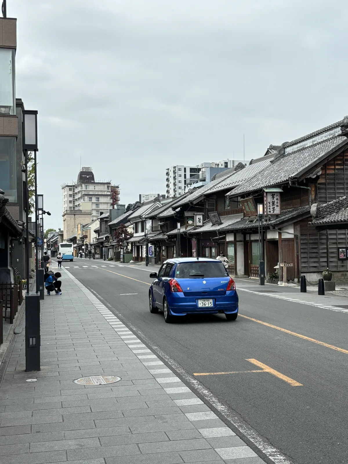 從路中央往北看。兩側是連續的蔵造り町屋，黑瓦、厚實的屋脊、二樓開著小窗。路面畫著黃色中線，一輛藍色的車經過，遠方的天際線混著現代的高樓。人行道上只有兩三個人。