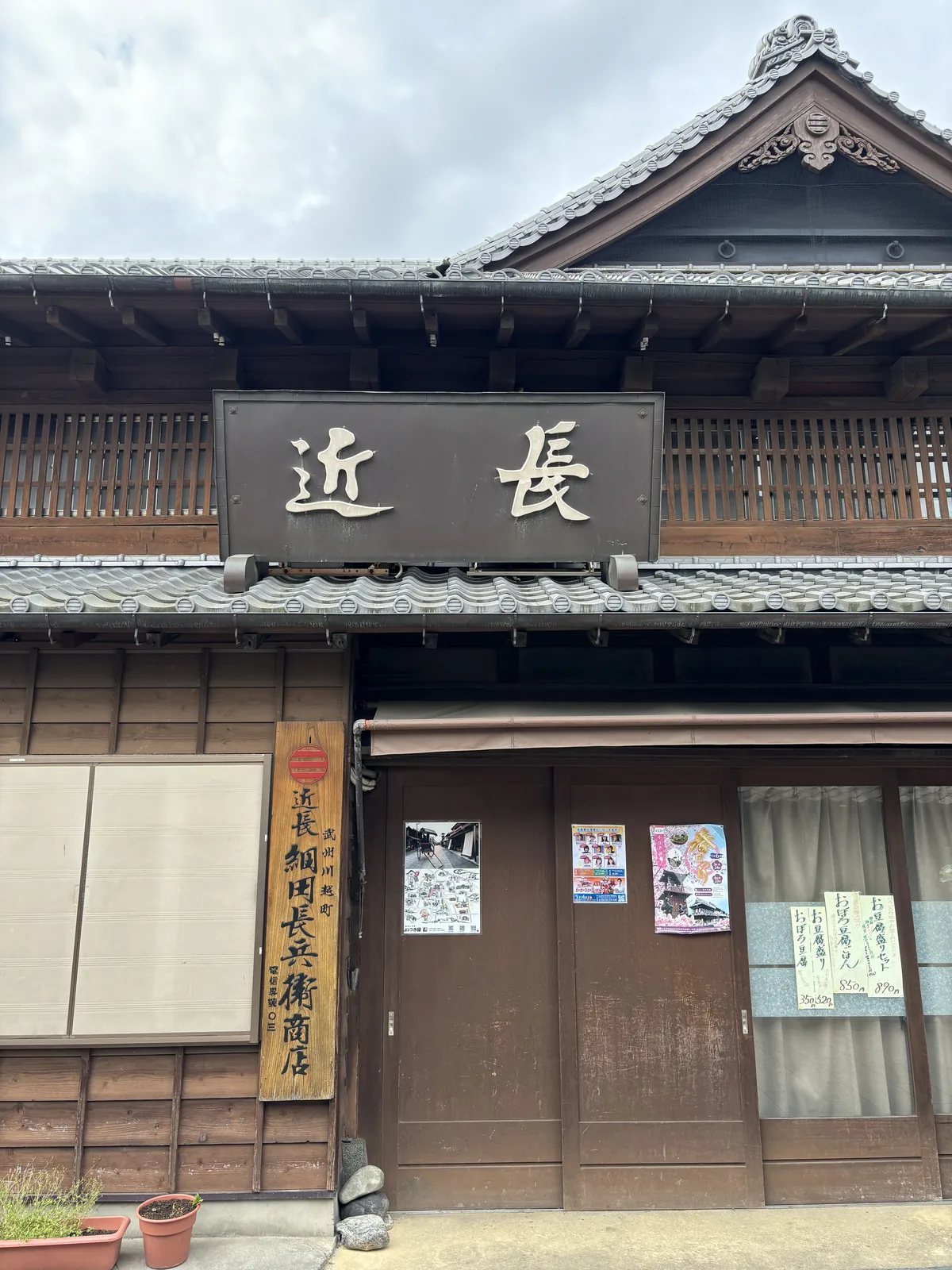 木造商家的二樓，深褐的木格子與白色的漆喰。屋簷下懸著一面大木匾，黑底金字由右至左寫「近長」。下方垂著米白色的暖簾，右側木柱上掛一塊直書的木牌與幾張貼紙。