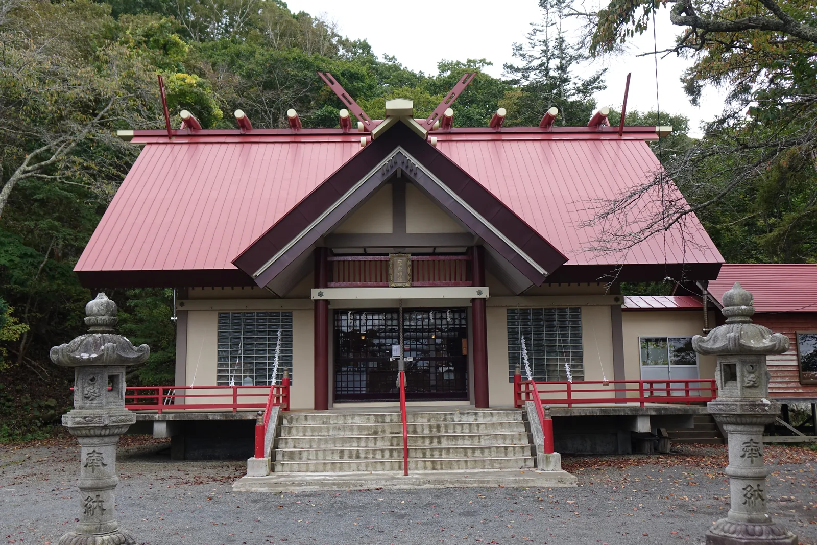 厚岸神社的拜殿，紅色鐵板豎葺的兩坡屋頂，屋脊兩端與正中各立千木、其間分列鰹木，前面另出一個向拜破風，前景一對石燈籠刻「奉納」