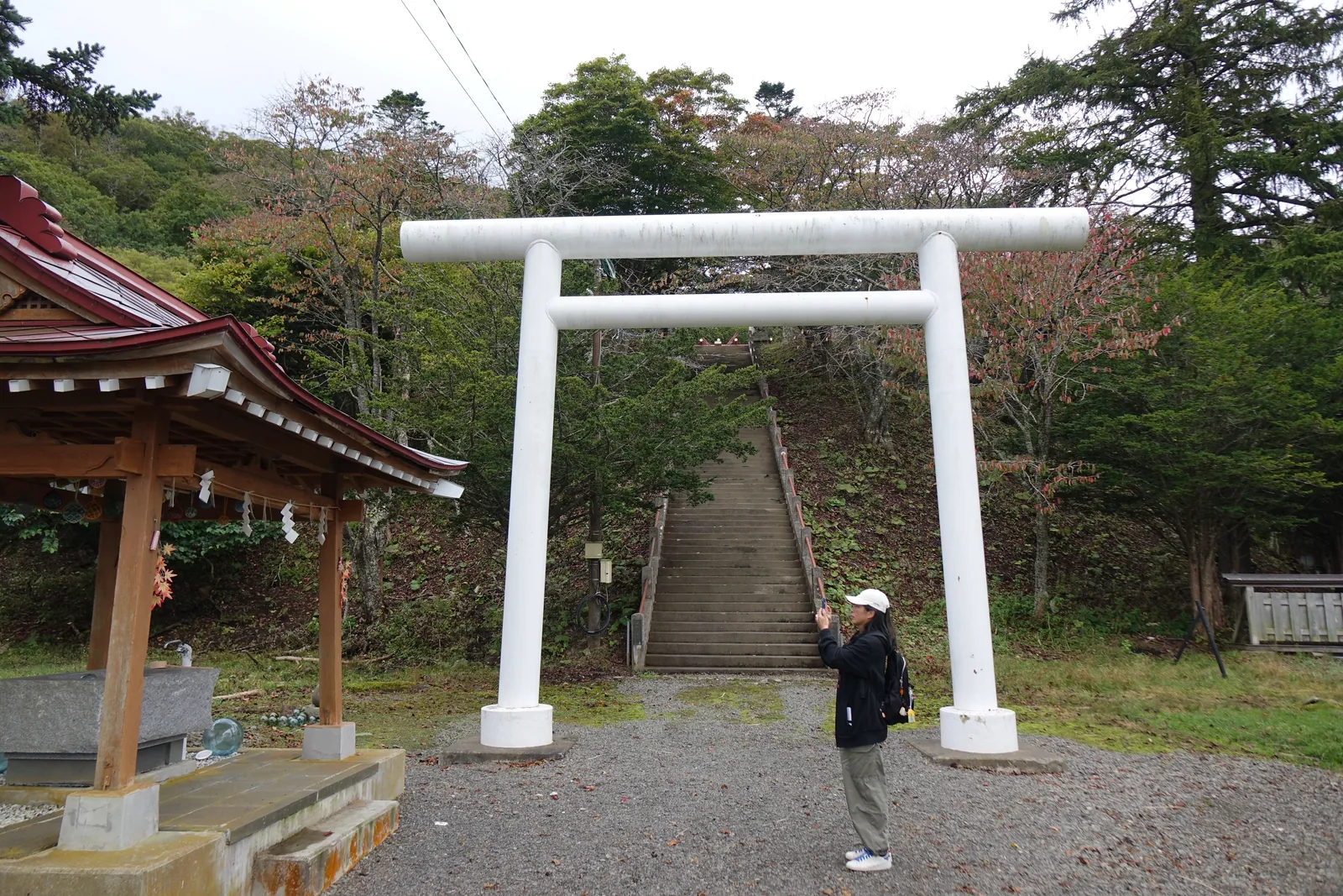 厚岸神社的白色一之鳥居與背後又直又陡的石段，左邊是木構紅屋頂的手水舍，水盤石台旁擺著幾顆綠色與藍色的玻璃浮球