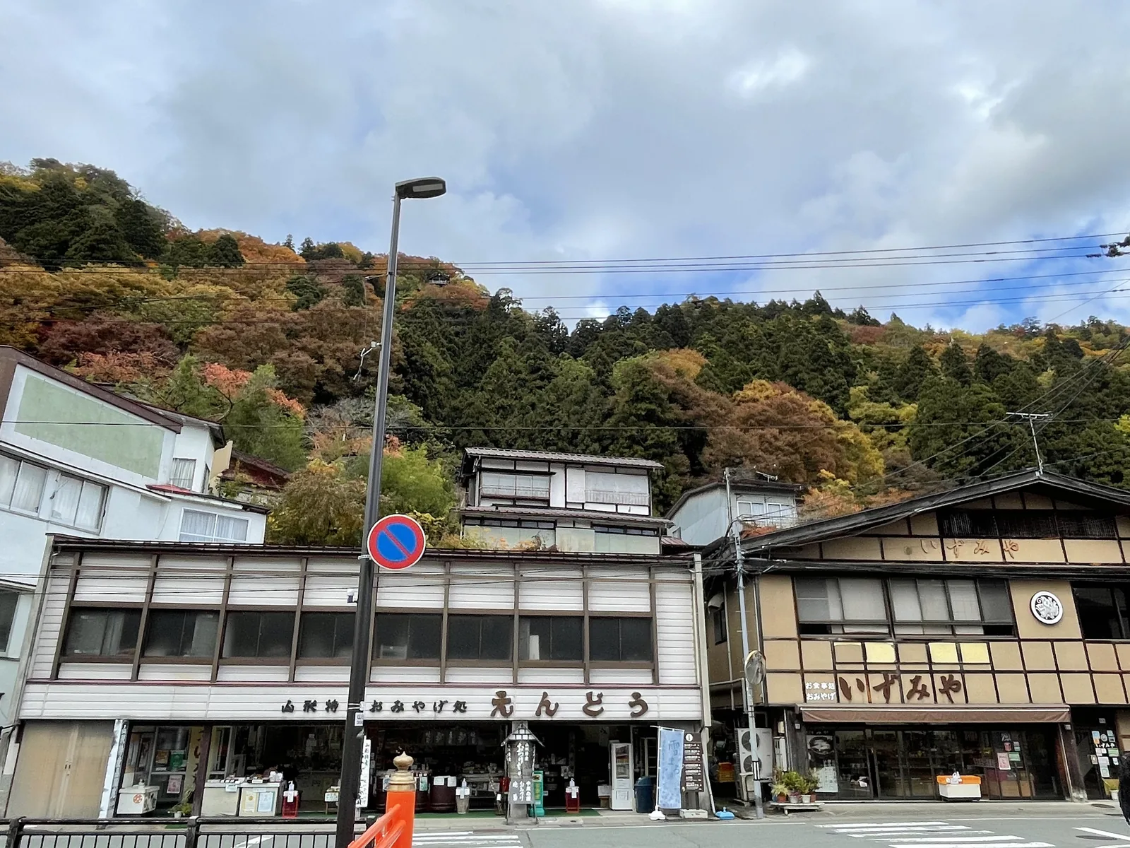 兩層樓的店面連成一排，招牌寫「山形特產　おみやげ処　えんどう」（山形特產土產店　遠藤）與「お食事処・おみやげ　いずみや」（餐館兼土產店　和泉屋）。房子背後的山坡直接立起來，坡頂的樹已經紅了；崖上一個小小的黑點就是五大堂。