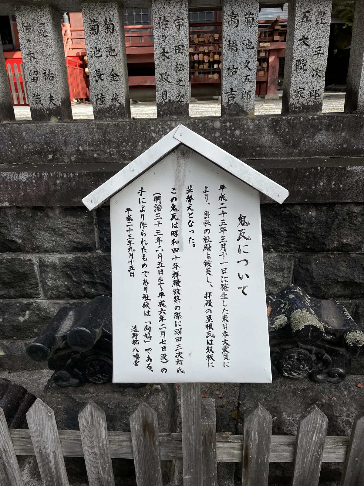 手寫的說明牌，立在卸下的鬼瓦前面。內容是：平成二十三年三月十一日的東日本大震災使社殿受災，拜殿的屋瓦改葺為鐵板；這些鬼瓦是拜殿改築時由沼田三次助所作，社紋為「向鳩」。落款日期平成二十三年九月十五日。牌後那排刻著捐納者姓名的石柱是玉垣。