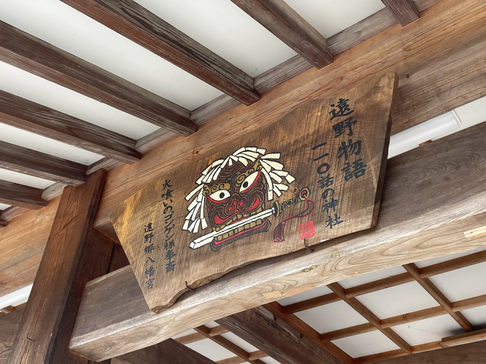 掛在境內一處屋簷下的木札，字面意思是「《遠野物語》第一一〇話的神社」。上面畫著一張咬住刀的獅子頭，兩側寫「火喰いのゴンゲン様奉齋」（供奉吃火的權現樣）與「遠野鄉八幡宮」。柳田記的那尊會吃火的權現樣，一年只有一月十五日、只在拜殿才拜得到。