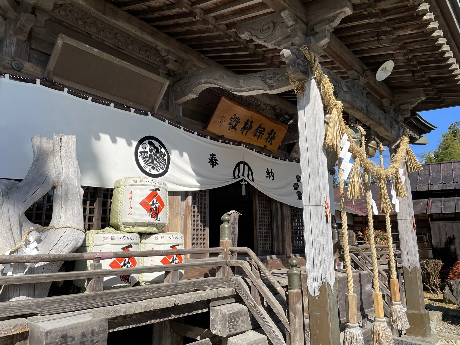 素木不上漆，木鼻與蟇股刻雲形，正面掛白幕、幕上是黑色圓形社紋，中央木製扁額刻「秋保神社」。階前堆著奉納的酒樽，注連繩垂著稻藁與紙垂。左側那段泛白的木頭是長床裡的おもかるの木。