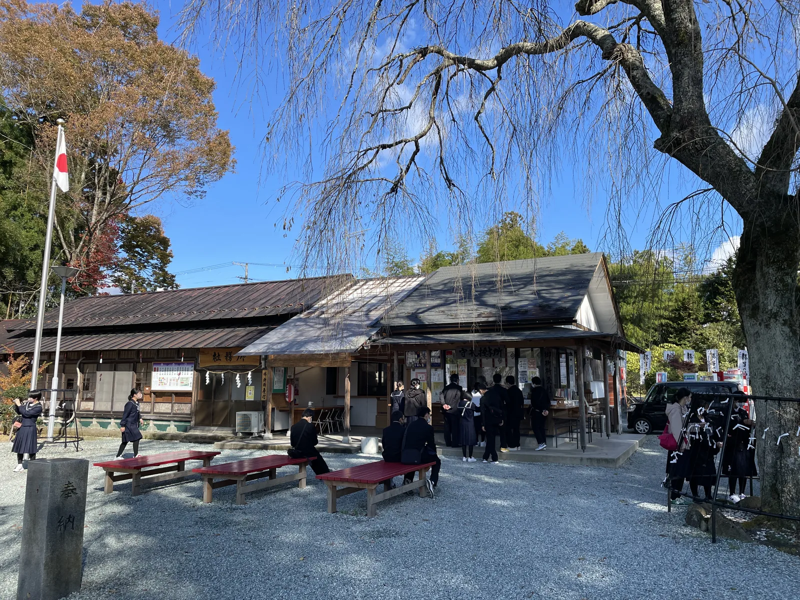 左邊掛「社務所」木牌的長屋是舊建物，右邊是守札授与所。那天早上十點，一群穿制服的學生排在授与所窗口前。前景的枝垂櫻已經落盡，只剩枝條垂下來。