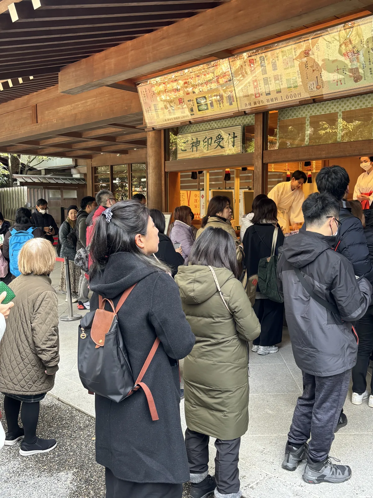 授与所的窗口前排著隊。木牌題「ご神印受付」，上方一整排燈箱列著授与品的照片與價格，其中一行寫「各種　御守り　初穂料 1,000円」（各式御守，初穗料一千圓）。窗口內側站著兩位穿淺色千早（ちはや chihaya，巫女在神事時罩在最外層的薄衣）的巫女，外側是二十來個等待的人。