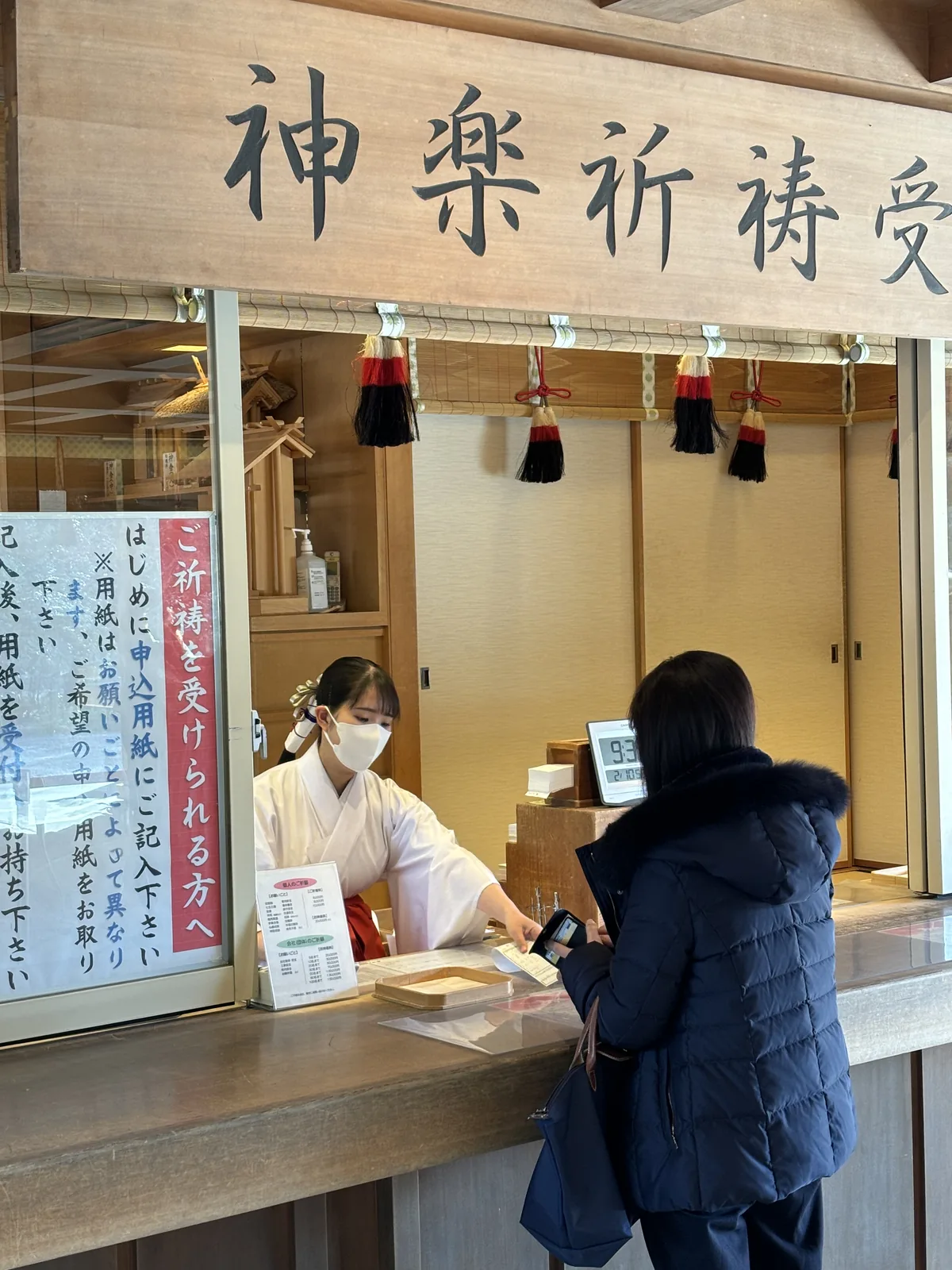 另一個窗口。杉木的橫牌題「神楽祈祷受付」，簷下垂著紅白黑三色的房飾。窗內一位巫女隔著櫃臺把申込用紙遞出來，左側的白色告示寫「ご祈祷を受けられる方へ　はじめに申込用紙にご記入下さい」——要受祈禱的人，請先填寫申請單。