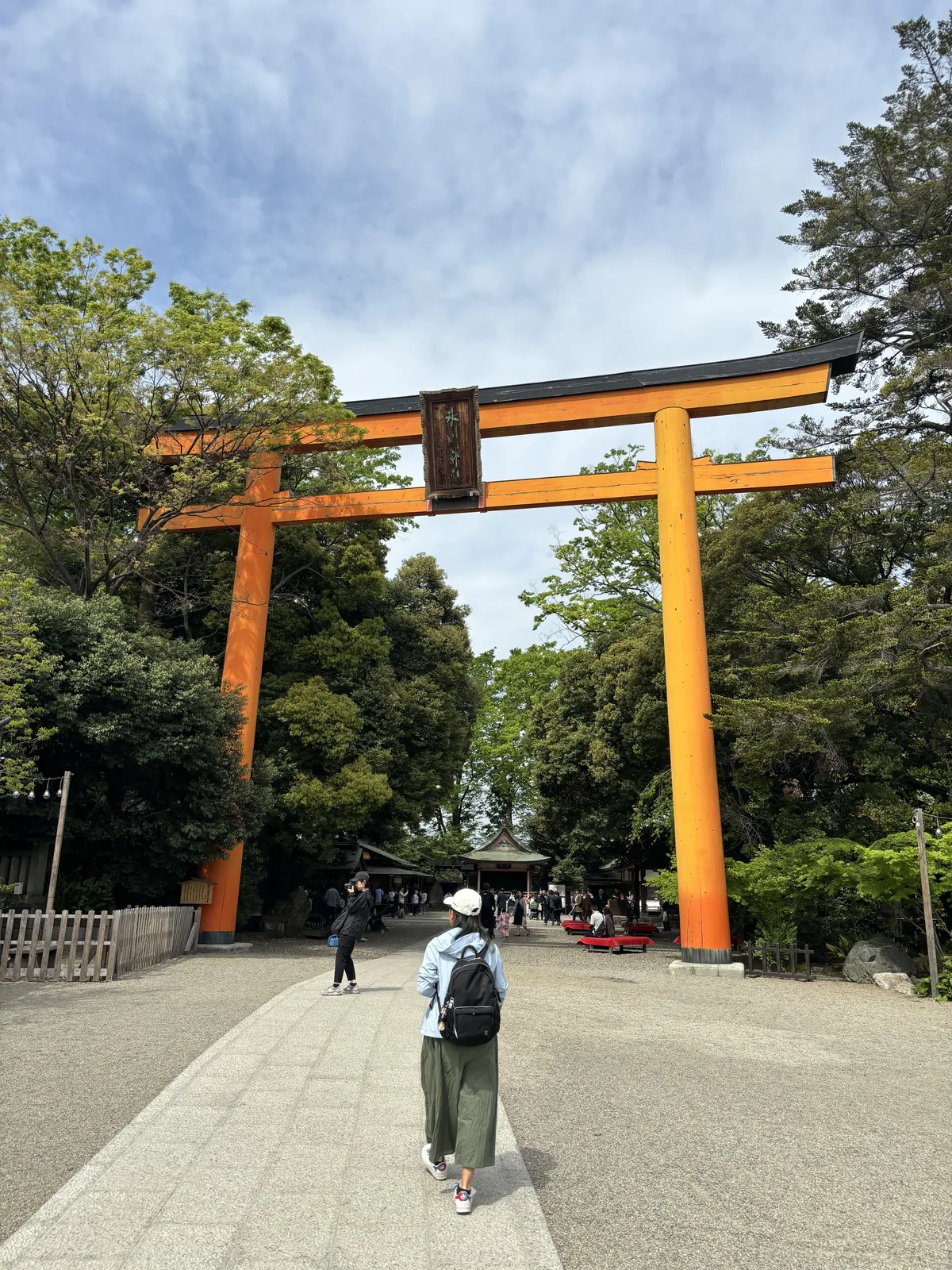 東參道口的大鳥居。朱漆的木造明神鳥居（みょうじんとりい，myōjin torii，笠木兩端上翹、下加島木的鳥居形式），兩根柱子粗到一個人抱不住，笠木的兩端微微翹起，中央懸一面深色的扁額。鳥居後方是碎石鋪面的參道與兩側的常綠樹叢，一個背著背包的人正走進去。上午九點五十三分，天是薄雲。