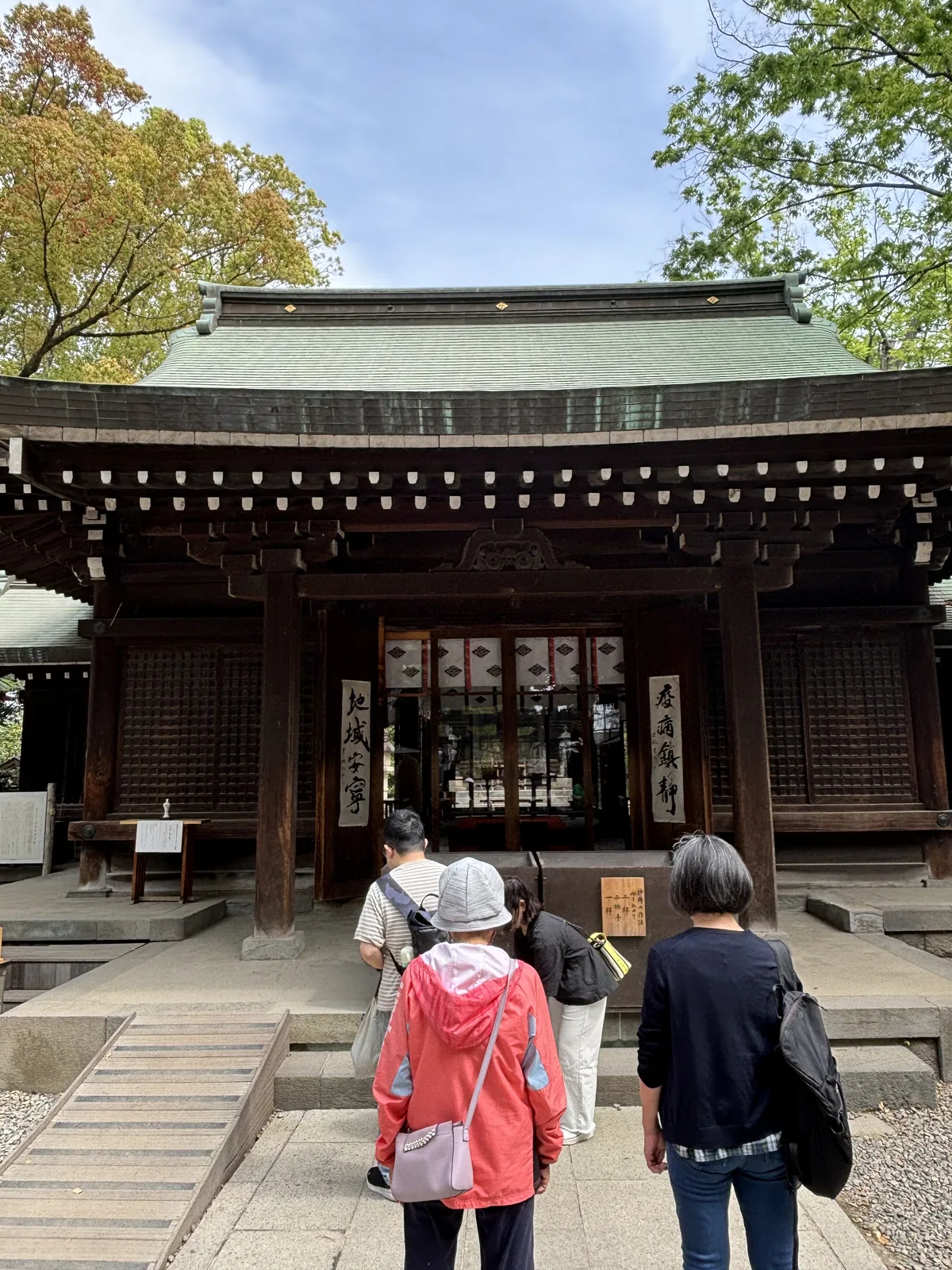 四坡頂上方接一段兩坡頂的屋面，鋪著深色的金屬板，正面挑出一段弧形起伏的破風。簷下一排斗栱與雕刻，正面三面白底的大板並排懸著。階前是一道木階與一道緩坡，三個人正走上去。左右是深綠的樹。
