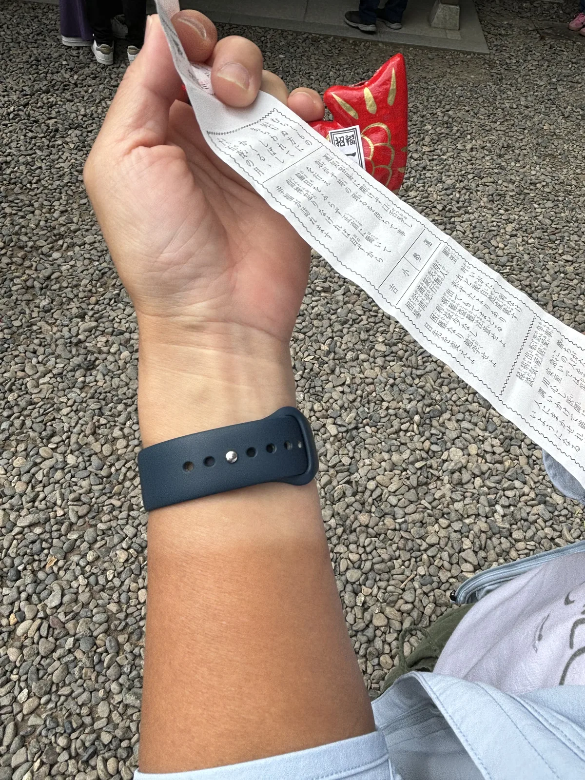 一隻手舉著一張長條的白紙，上面直排印著籤文；另一隻手拎著一尾紅色的鯛，鯛的側面印著一條白色的細帶。背景是境內的碎石地與一雙運動鞋。