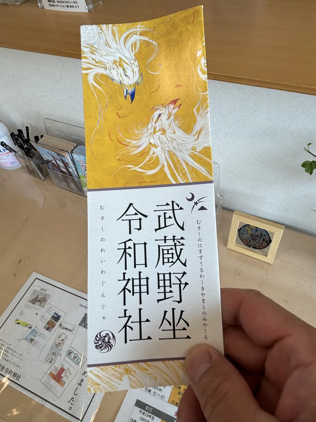 一隻手拿著一份直式摺頁。上半是金地上一龍一鳳交纏的白描，下半白底直書「武蔵野坐令和神社」，右旁小字註「むさしのにますうるわしきやまとのみやしろ」，左旁註讀音「むさしのれいわじんじゃ」，社名下方是一枚圓形的鳳凰社紋。背景是授与所的木櫃檯，左側有摺頁架與筆筒，右側立著一塊嵌著黑色圓形紀念章的小木框。