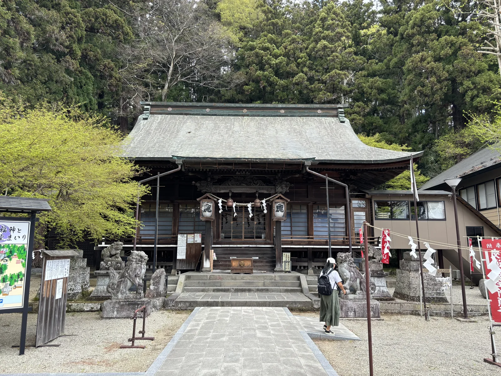 從碎石廣場正面看拜殿。上段兩坡、下段四坡的屋面鋪著泛出銅綠的金屬板，四角的簷端微微翹起。簷下一整面雕刻，中央垂著兩具寫「鹿」字的木造釣燈籠（つりどうろう，tsuridōrō，懸吊式的燈籠）與一條注連繩（しめなわ，shimenawa，界分神域的稻草繩）。階下左右各兩隻石造的狛犬（こまいぬ，komainu，鎮守在神社入口的一對石獸），一前一後坐在高低不同的石台上。左側立著一塊帶小屋頂的手繪境內圖案內板，右側是紅底白字的幟旗。背後整片是深綠的杉林，坡度往上——那是甕森。上午十點五十分。