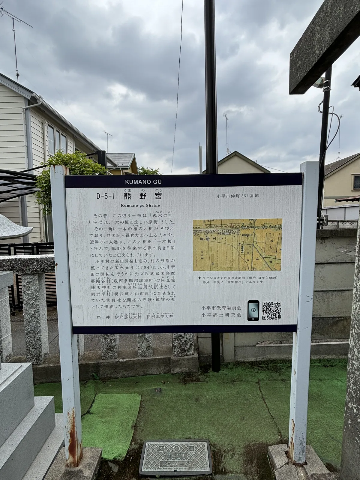 白底的說明板立在兩根鐵柱上，頂端一條深色橫帶寫「KUMANO GŪ」。板面左側是直排的日文說明，右上角標「D-5-1 熊野宮／Kumano-gu Shrine」與「小平市仲町 361 番地」，最下一行寫祭神兩位的名字。右側貼著一張泛黃的舊地圖，下方註明是明治十三年（一八八〇）的法式彩色版迅速測圖（明治初年以法國式測量法趕製的簡易地形圖）局部，中央標著「熊野神社」。板後是民宅、電線與陰天。