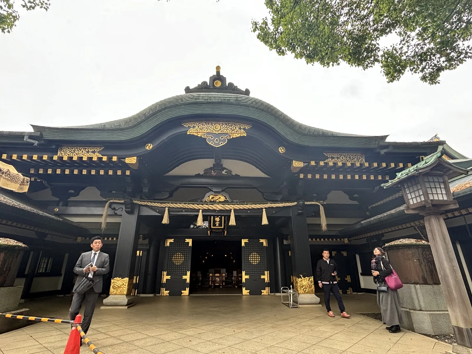 拜殿正面。柱、樑與門扇全部漆成黑色，上面釘滿金色的金屬件。正面挑出一段弧形起伏的破風（はふ，hafu，屋頂山牆邊緣那道斜向的構件），屋面鋪著氧化成青綠的銅板，正脊中央立一枚金色的菊紋，破風兩側各嵌一枚三巴紋。簷下的斗栱一格一格貼金，橫過一條稻草繩與四條白紙條，中央懸著寫「八幡宮」的匾額。兩扇門開著，殿內深處隱約可見陳設。左右各立一座大型的石燈籠，階前站著三個人。天是陰的。