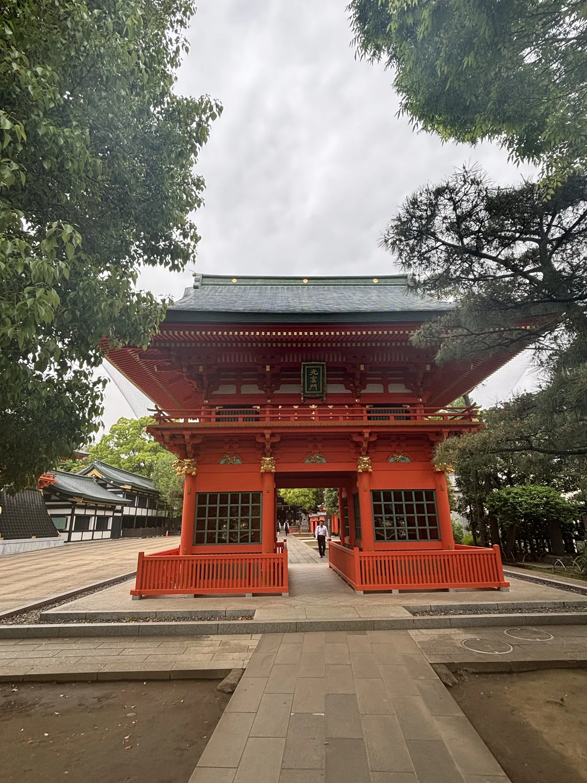 朱漆的二層樓門。上層的屋面鋪青綠的銅板，四角起翹；二層中央掛一面綠底金字的匾額，寫「光寮門」。下層左右各有一扇綠色格子窗，窗後奉安著神像。門前圍著朱漆的矮欄，地面鋪石板。左後方是白牆與青綠屋頂的建物，右側是黑松。一個人正從門洞裡走出來。