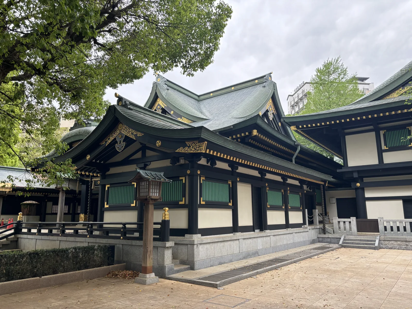 一棟黑柱、白牆、青綠銅板屋頂的建物，正面一排木格窗，屋簷下懸著木製的燈。屋頂的四角起翹，脊端有金色的裝飾。前方是石板鋪面與一道低矮的欄杆，右側可見另一棟建物的白牆。