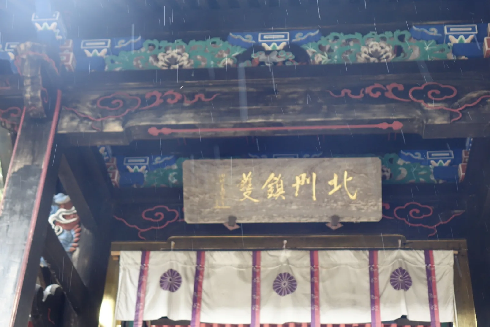 仰角看中門。上方是極彩色的斗栱：藍地、紅色的雲紋、綠白相間的牡丹，柱頭伸出彩繪的獸形木鼻。中央懸著一塊木匾，金字橫書「北門鎮護」，左端有一行細小的落款——解析度不足以判讀。匾下垂著一幅白幕，幕面用紫線分成數格，每格中央一個紫色的十六瓣菊紋。畫面上同樣有雨絲。