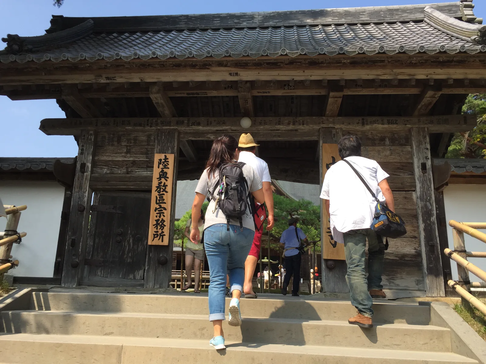 中尊寺本堂前的表門：四柱一列、主柱在前的門形式，本瓦葺的屋頂，兩側是白壁與竹籬，門洞左側掛著寫「陸奧教區宗務所」的長木牌，參拜者正踏石階進門