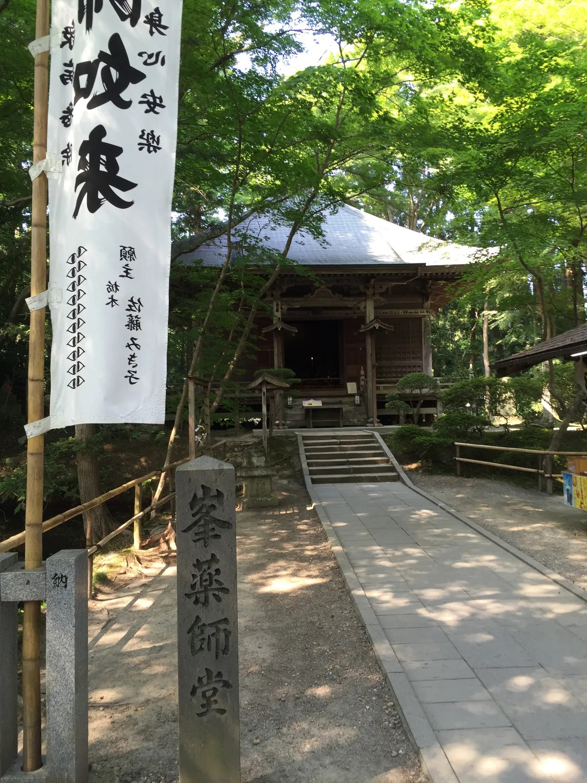 峯藥師堂前：白色的幟旗寫著「拔苦與樂」，旁邊立著刻有堂名的石柱，石段往上通到樹蔭下的小堂