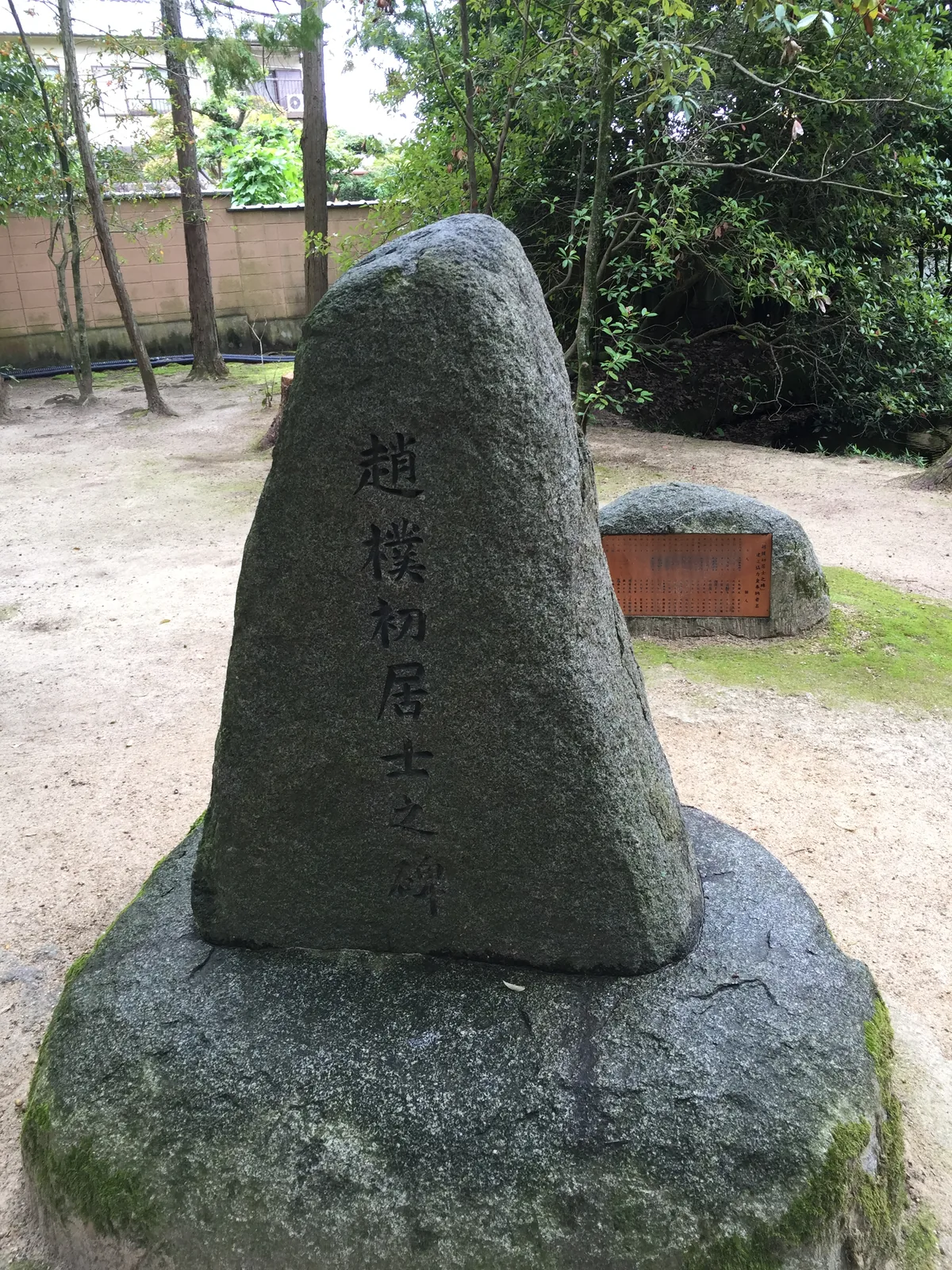 「趙樸初居士之碑」：一塊三角形的自然石立在方形石座上，正面刻著六個大字，後方的地上另有一塊帶銅板說明的矮石，四周是砂地與樹