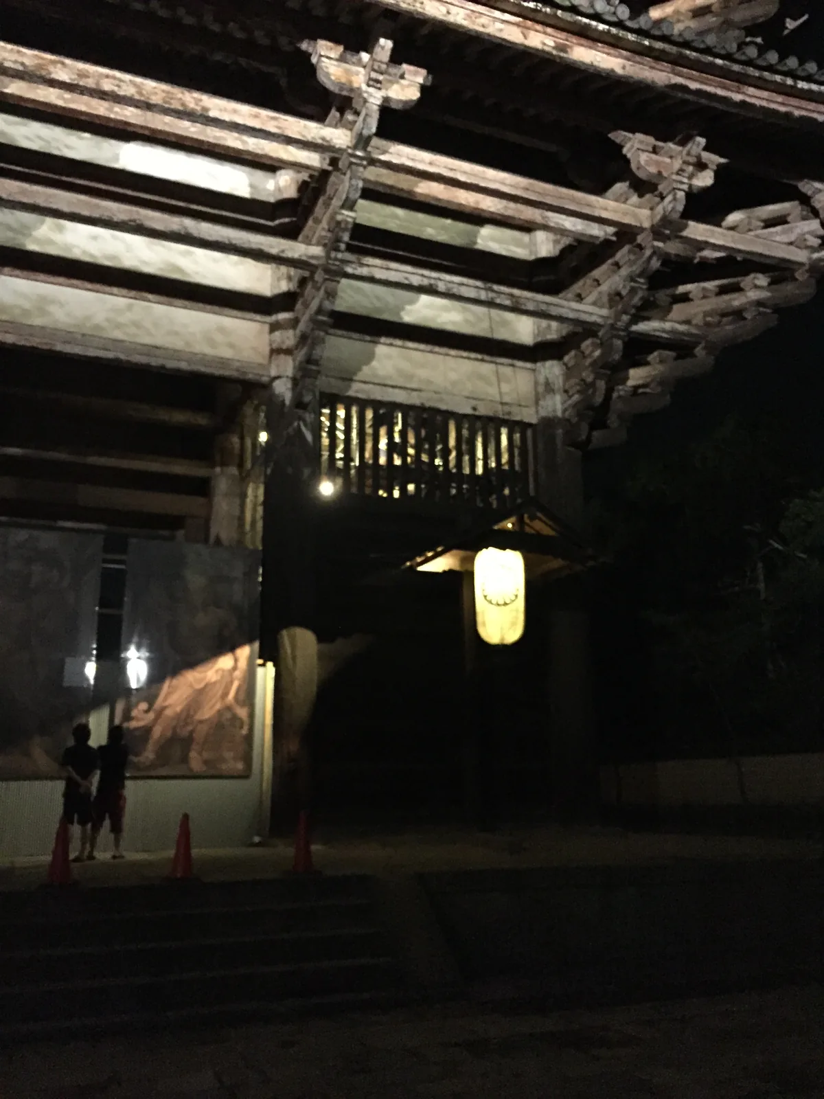 仰看南大門東南角的簷下：柱身上一層層插出的橫材在燈下數得一清二楚；左側一面圍板擋住其中一間，板上印著兩尊力士像的照片，兩個人站在板前看