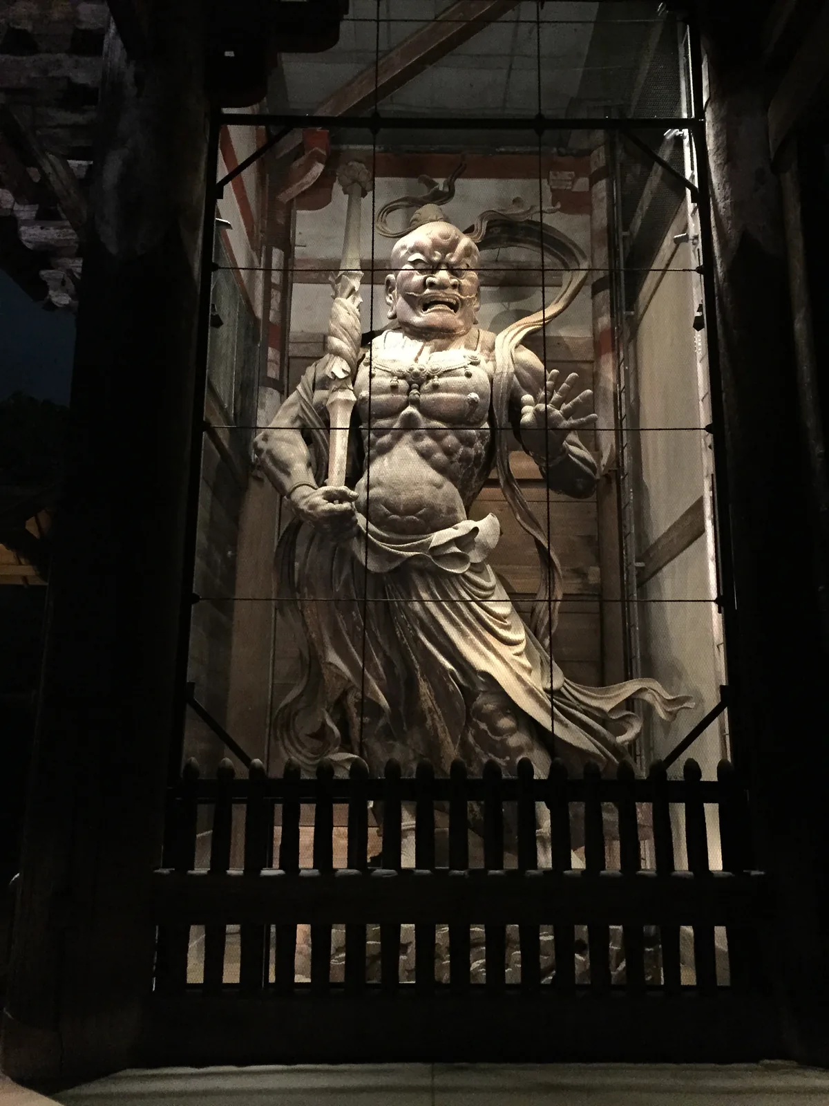 南大門內張著口的金剛力士像：一手握杵、一手張開，衣帶往兩側飛起；像前是黑色的木柵與金屬網，像後可以看見紅白相間的鷹架