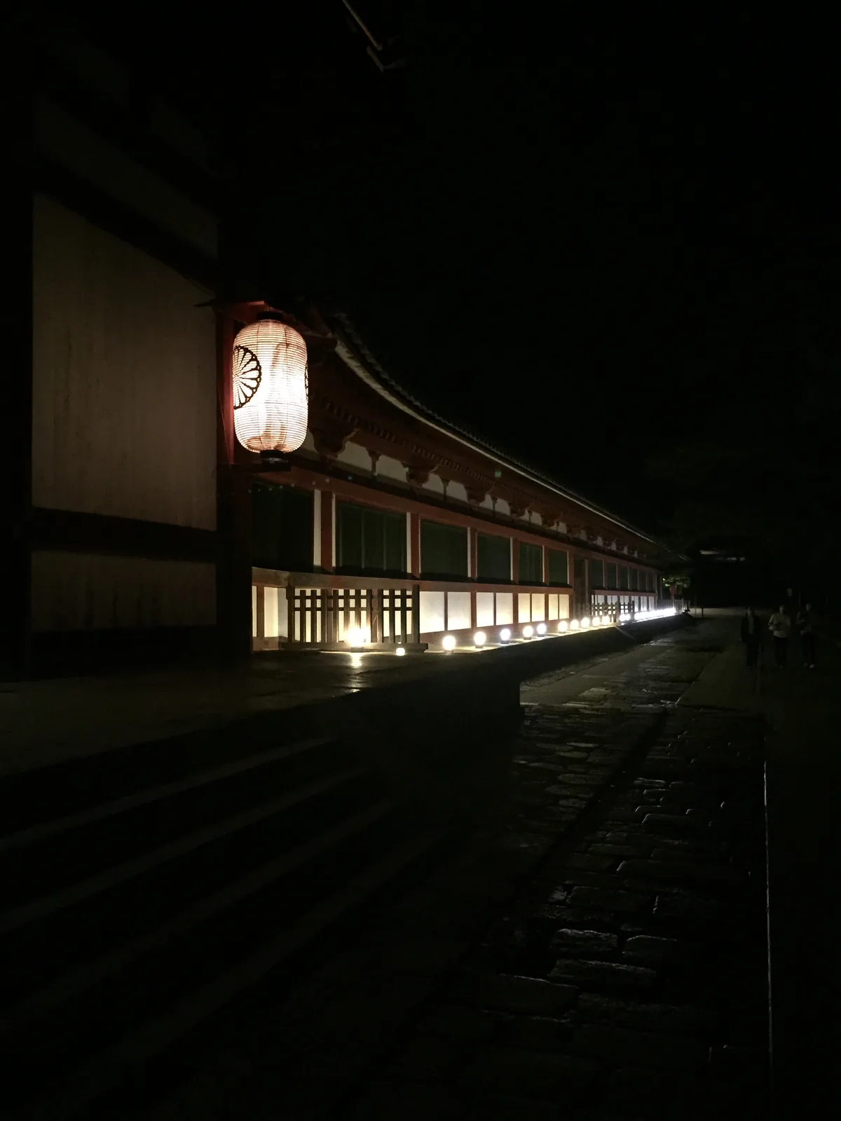 東大寺中門與往東延伸的迴廊夜景：一盞大紙燈籠掛在屋簷下，迴廊的下緣被地燈打亮成一條水平的光帶，屋簷以上全黑，右下是濕的石板路
