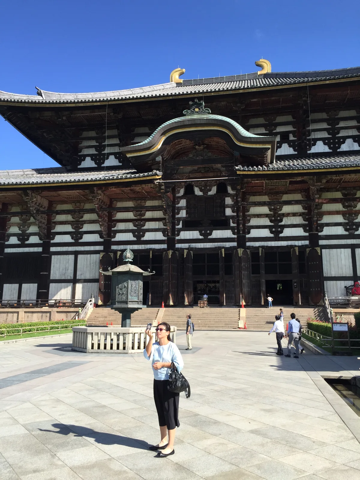 東大寺大佛殿正面的白天：巨大的斜坡屋頂、屋脊兩端的金色飾物、中央一座弓形的裝飾山牆與其上的大窗；殿前石砌的台上立著一座八角形的銅燈籠，前方是寬闊的石鋪廣場與一位遊客
