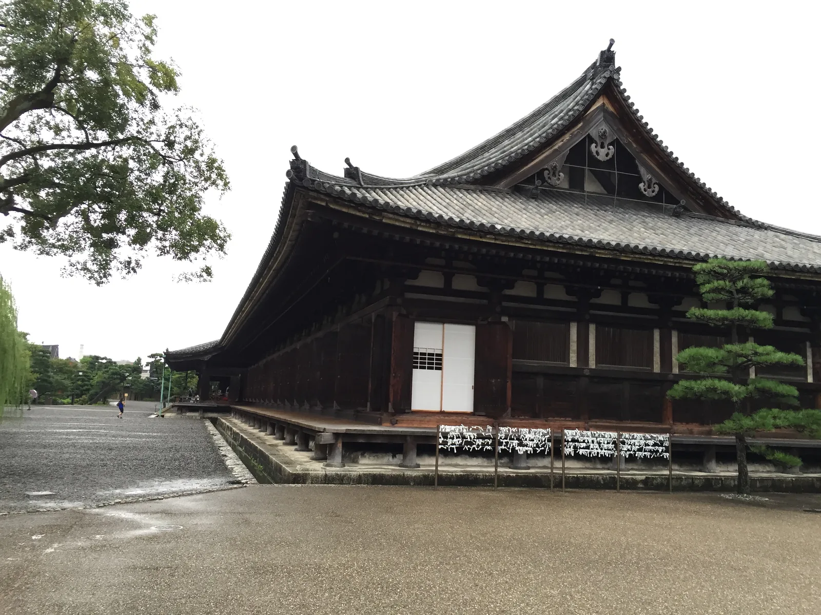 三十三間堂本堂的北端，自東北看：右邊是一面懸山的山牆，屋脊兩端立著飾物；左邊那一面往南退去的長側面看不到盡頭，白色的板門與深色的木構交替，簷下欄杆上綁滿白色的籤，地面是濕的碎石