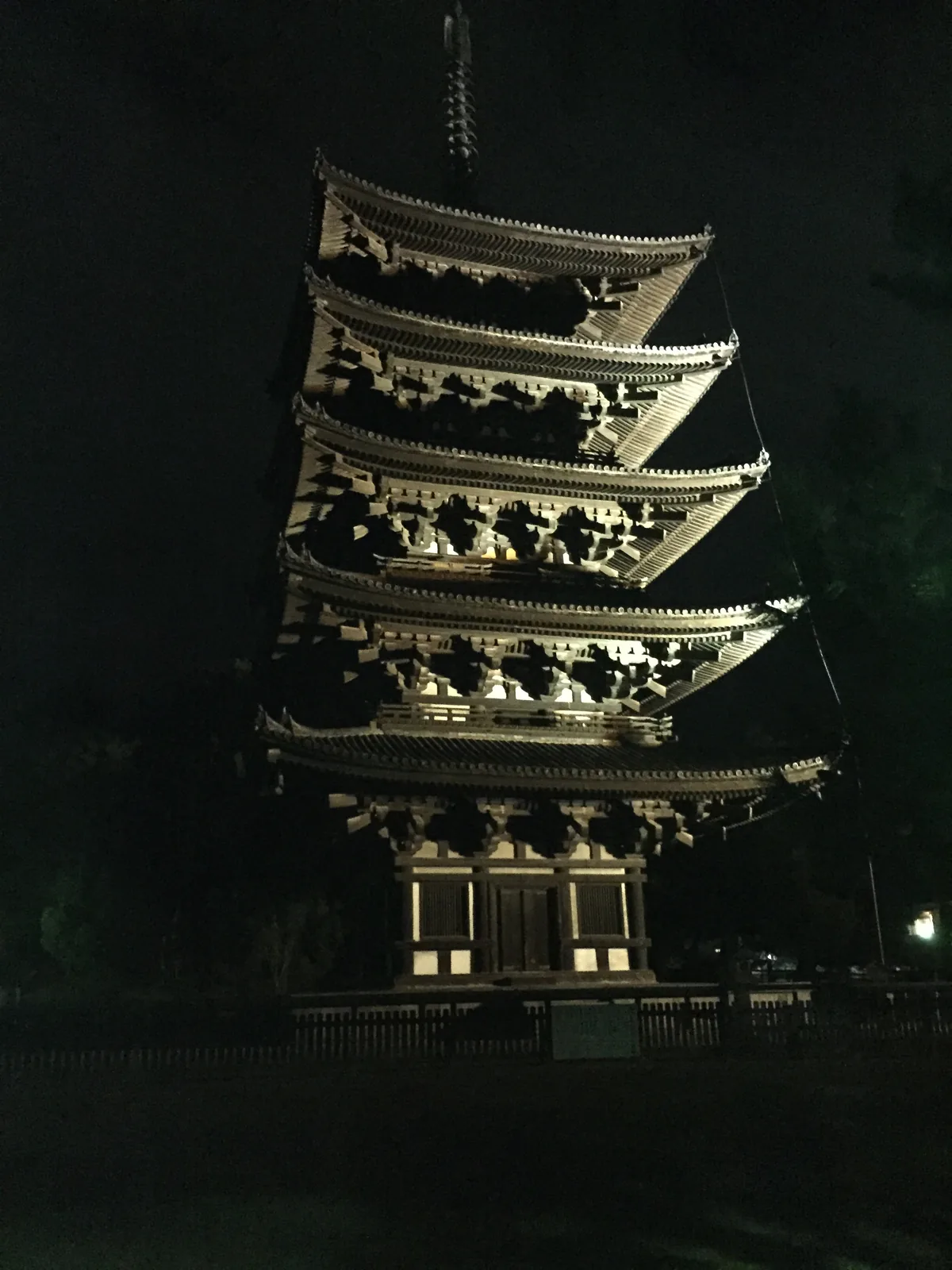 興福寺五重塔的夜間照明：塔身在暗處，只有五層的軒與初層的板扉被下方的燈打亮，三手先斗栱（みてさきときょう，向外挑出三層的斗栱）的每一層都看得出挑出的深度，塔頂的相輪（そうりん，塔頂的金屬柱狀構件）伸進全黑的天空，塔下是木製的玉垣與一面說明板