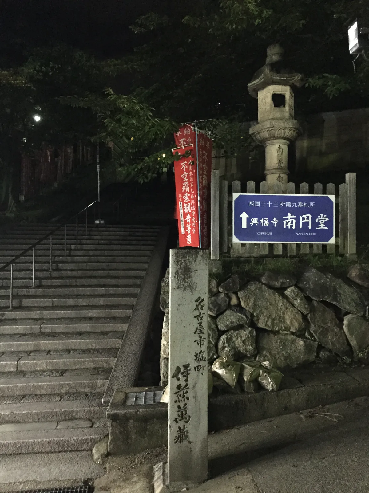 五十二段下的路口：藍色案內板寫著「西国三十三所第九番札所　↑興福寺　南円堂」，旁邊一面紅幟寫「奉納　西国札所第九番　興福寺南円堂　不空羂索観音菩薩」，右側石燈籠的竿上刻著堂名，正中一根石柱刻「名古屋市塩町　伊藤萬藏」，左側是往上的寬石階