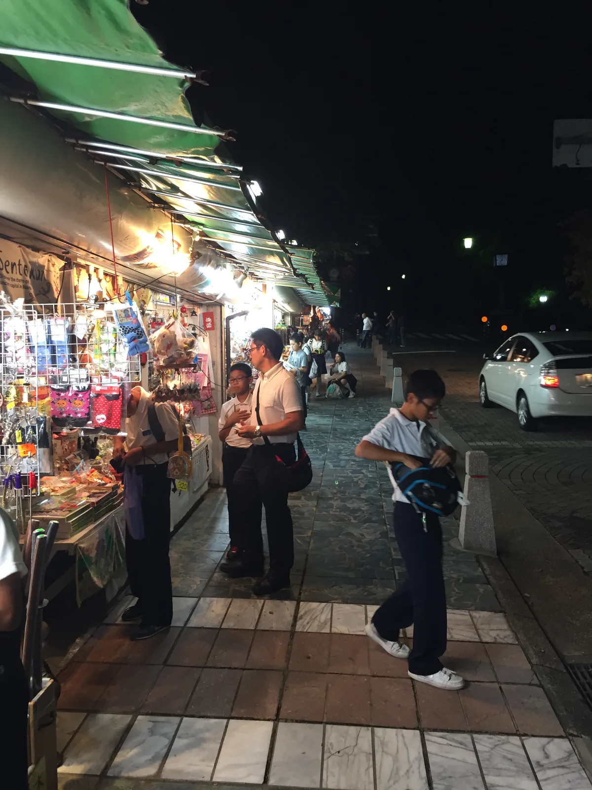 猿澤池北岸的露店：綠色遮陽篷下掛滿鑰匙圈與玩偶，一群穿白襯衫深藍長褲的中學生正在挑東西，人行道外側就是車道