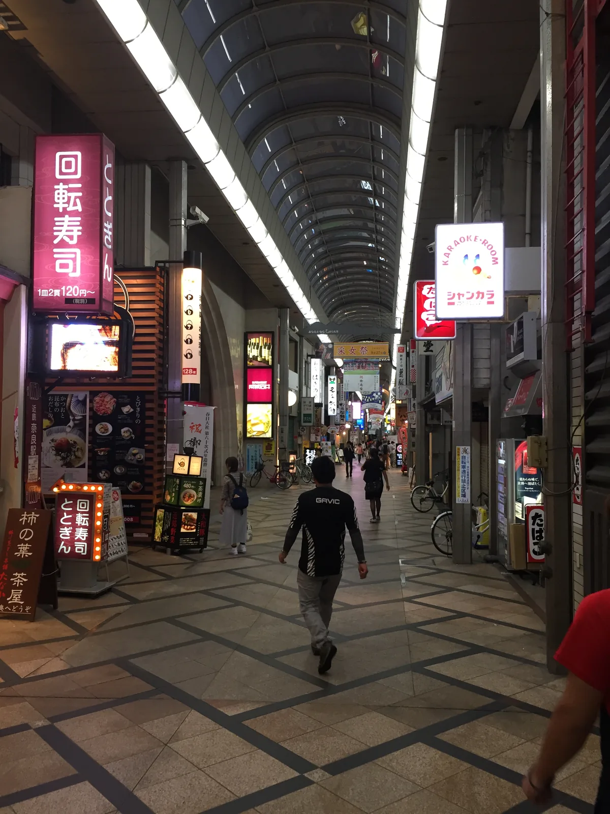 東向商店街的拱廊：兩側是回轉壽司、藥妝店與土產舖，天花的燈把整條路照得比外面亮，幾名行人往深處走