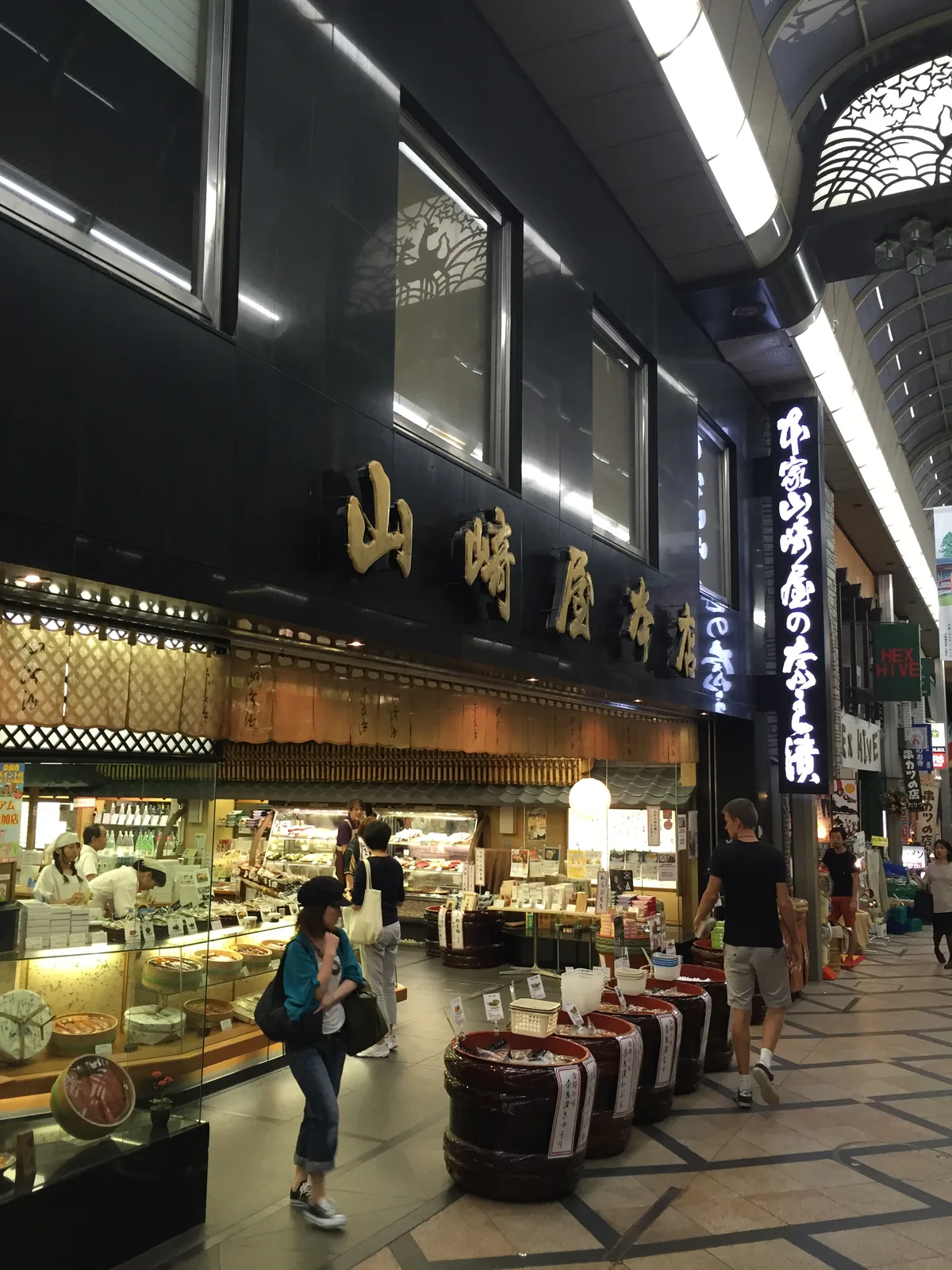 商店街裡的和菓子老舖：黑色的木造店面，簷下掛著橫書招牌，玻璃櫃裡排著菓子與提籃，店內的燈亮著