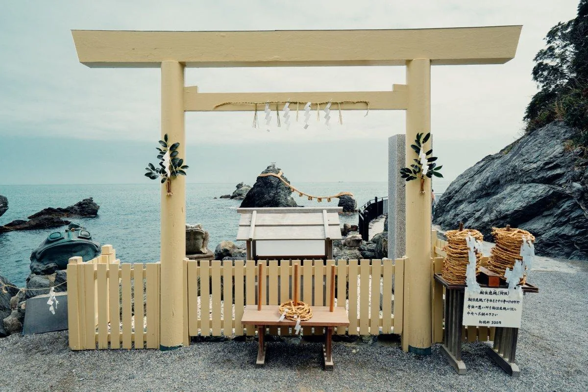 二見興玉神社的遙拜所：素木的鳥居正對著海，橫樑上掛著注連繩（しめなわ，隔開聖域的稻稈繩）與紙垂（しで，摺成鋸齒狀的白紙），兩根柱上各綁一束榊（さかき，神事用的常綠樹枝）；鳥居框住的正中央就是夫婦岩，大注連繩垂在兩岩之間，大岩頂上立著一座很小的鳥居，右側木台上疊著兩落輪注連繩