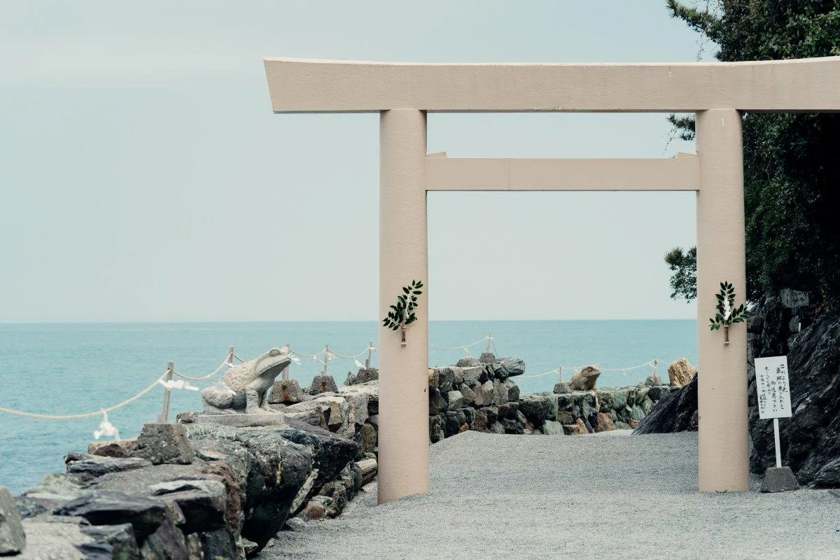 二見興玉神社參道突堤上的鳥居：素木、兩柱各綁一束榊，穿過去是沿海岸鋪碎石的參道，左右的石積護岸上各蹲著一尊石造的蛙，左側是拉在木樁之間的注連繩，再過去就是海