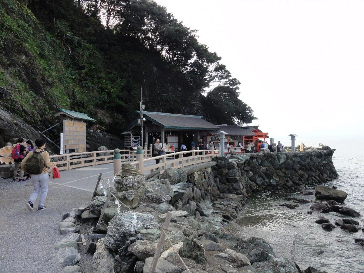 二見興玉神社沿海岸的參道：左側是石積的護岸與外海的岩礁，右側貼著山崖立著一列社殿與朱色的建物，參拜者沿著木欄杆往前走