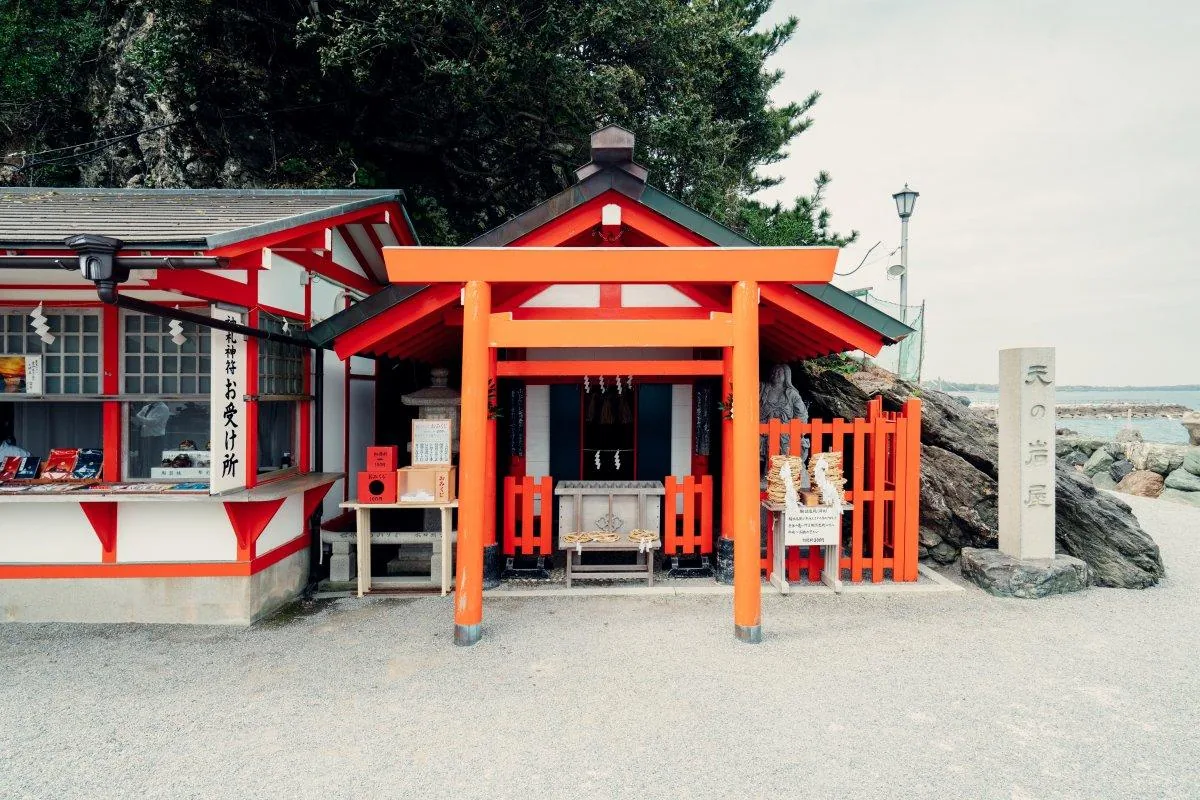 天之岩屋：朱色的鳥居與小社貼著岩壁建起來，右側立著刻「天の岩屋」的石標，左側是寫「御札神符　お受け所」的授與所，社前的木架上疊著整落的輪注連繩