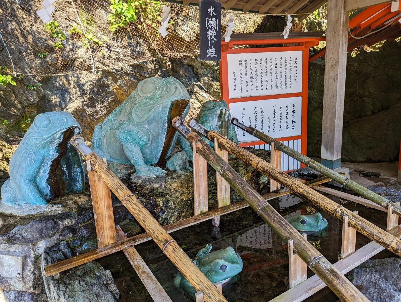 二見興玉神社的手水舍：三尊青綠色的大蛙蹲在岩上，水從牠們身下流進水槽，竹製的柄杓橫架在上面，水裡還沉著兩尊小蛙；上方黑底的木牌寫「水（願）授け蛙」——授水授願的蛙——，右側兩塊白底說明板，一塊題「二見蛙について」並落款「二見興玉神社」，另一塊寫「手水の仕方」的四個步驟