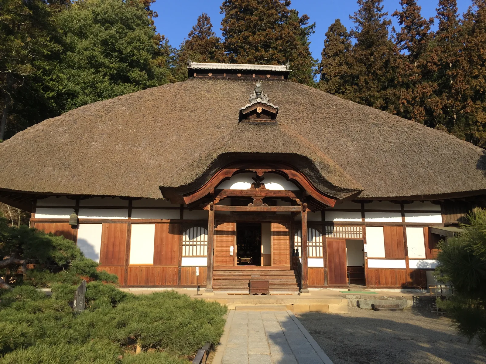 常樂寺本堂正面全景：寄棟造（よせむねづくり，四面斜坡的屋頂）的茅葺（かやぶき，茅草屋頂）大屋頂壓得很低，正面中央的茅被刳成一道唐破風（からはふ，中央拱高的曲線山牆）的弧，弧內露出木造的破風板，屋脊上另架一座瓦葺的小越屋根，早晨的光從東南方打上正面